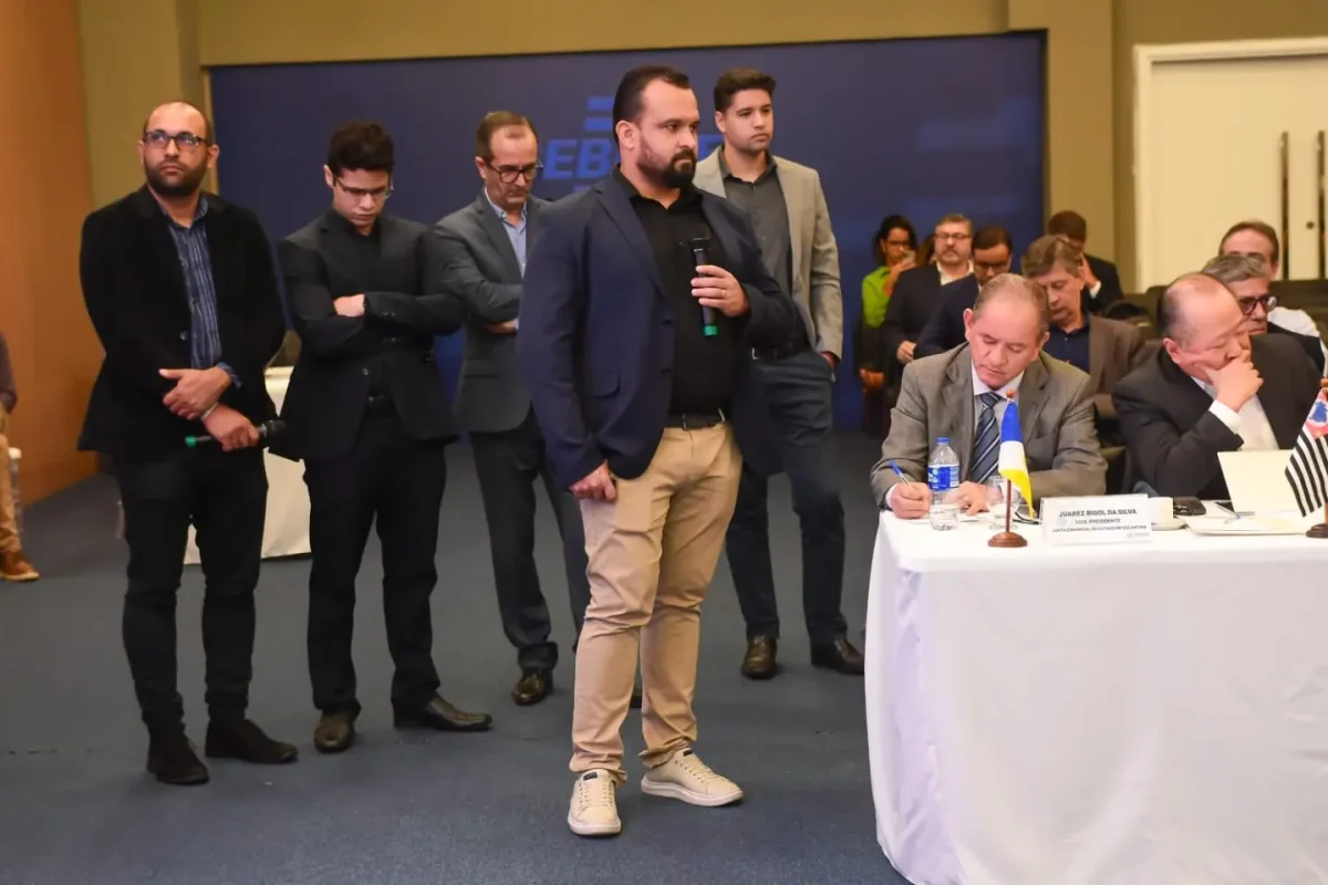 Aiuá Reis com microfone durante um encontro, entre participantes e a mesa do evento.